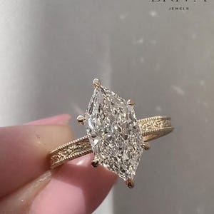 Puede incluir: Un anillo de compromiso de oro con un gran diamante talla marquesa. La banda tiene un diseño grabado. El anillo es de la marca BRIVA JEWELS.