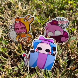 May include: Three colorful stickers featuring cartoon characters. One sticker shows a character in a yellow costume, another shows a pink character with text, and the third shows a character in a blue gift box. The stickers are on a bed of green grass.