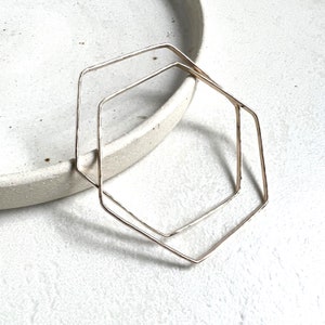 May include: Two gold hexagon shaped hoops, one slightly larger than the other, are stacked on top of each other. The hoops are made of thin metal and have a slightly hammered texture.