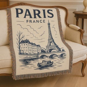 May include: Cream-colored throw blanket featuring a navy blue illustration of the Eiffel Tower, buildings, and a boat on a river, with the text "PARIS FRANCE". The blanket has a fringed edge.