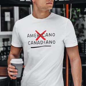 May include: White t-shirt with the text "AMERICANO" crossed out in red and "CANADIANO" below. The person is holding a grey coffee cup and wearing glasses and jeans. The background includes a black awning and outdoor seating.