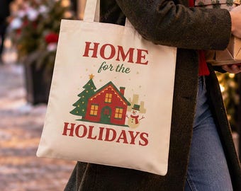 May include: A cream-colored canvas tote bag featuring the red text "HOME for the HOLIDAYS" above a graphic of a red house, a green Christmas tree, and a snowman. The bag is carried by a person wearing a dark green coat and a white knit beanie.