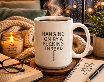 May include: A white ceramic coffee mug featuring the black printed text HANGING ON BY A FUCKING THREAD with a small thread graphic dangling from the last word. The mug has a smooth finish, a curved handle, and is filled with a dark liquid emitting steam.