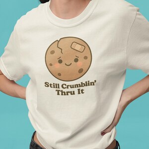 May include: Off-white t-shirt with a cartoon cookie design. The cookie has a bandage, a crack, and chocolate chips. The text below the cookie reads "Still Crumblin' Thru It".