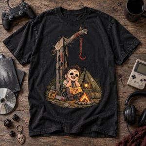 May include: A black short-sleeve t-shirt featuring a graphic illustration of a cartoon character with a stitched mask sitting by a campfire. The scene includes a tent, a chainsaw, a wooden post with a hanging hook, and a lantern.