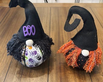 May include: Two Halloween gnome decorations. One gnome has a black hat with the word "BOO" in purple, a gray beard, and a patterned body with skulls. The other gnome has an orange beard and a black hat. Both have white noses.
