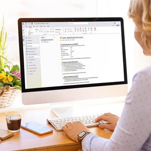 Op de afbeelding: Een persoon werkt op een desktopcomputer met een wit toetsenbord en een groot beeldscherm. Een kopje koffie, een notitieblok, een pen en een smartphone staan op het houten bureau. Een mand met bloemen staat in het raam.