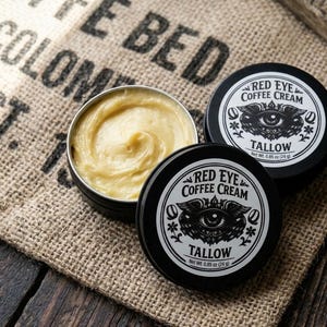 Könnte beinhalten: Zwei schwarze, runde Dosen mit Red Eye Coffee Cream Tallow, eine davon offen mit hellgelber Creme und eine geschlossen mit einem Etikett, das ein Augengrafik und den Text Net Wt. 0.85 oz (24 g) zeigt. Die Etiketten tragen den Produktnamen.