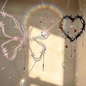 Op de afbeelding: Twee hangende decoraties: een roze vlinder en een zwart hart, beide gemaakt van prikkeldraad. De vlinder heeft bungelende kristallen en een ketting, terwijl het hart rode kralen en een zilveren ketting heeft. De stukken zijn opgehangen aan zilveren kettingen.