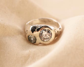 May include: A silver-toned ring with an irregular, textured band featuring two round, clear, faceted stones of different sizes set into the metal. The surface of the ring has a hammered, organic appearance with a polished finish around the stones.