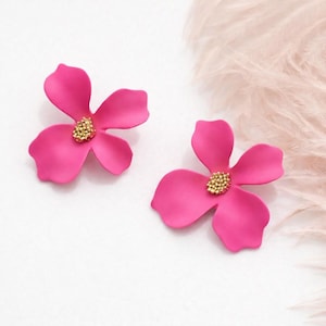Puede incluir: Dos pendientes en forma de flor rosa brillante con un centro dorado. Los pendientes están sobre un fondo blanco con un acento suave y plumoso. Los pétalos son lisos y curvos.