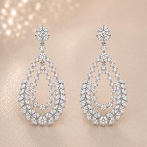 May include: A pair of silver-toned drop earrings featuring a teardrop-shaped frame encrusted with numerous small, clear, round and marquise-cut stones. Each earring hangs from a single round stone stud, creating a layered, openwork design.