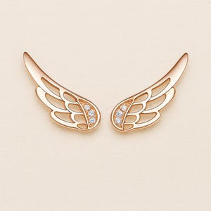 May include: A pair of rose gold wing-shaped earrings. Each earring features an openwork design with a cluster of small, clear gemstones near the base. The wings have a smooth, polished finish and are displayed against a soft, neutral background.