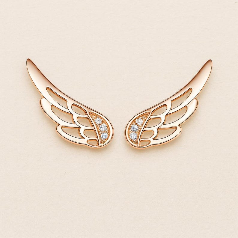 May include: A pair of rose gold wing-shaped earrings. Each earring features an openwork design with a cluster of small, clear gemstones near the base. The wings have a smooth, polished finish and are displayed against a soft, neutral background.