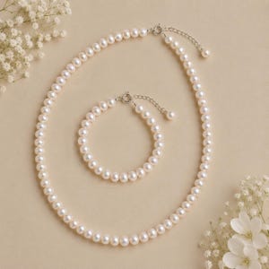 Puede incluir: Un collar de perlas cl&aacute;sico y un conjunto de pulseras a juego. La joyer&iacute;a presenta un hilo de perlas redondas, blanco crema, con un cierre plateado. El collar es m&aacute;s largo, mientras que la pulsera es m&aacute;s corta. El conjunto se muestra sobre un fondo neutro con flores blancas.