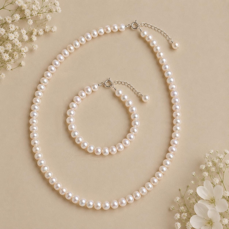 Puede incluir: Un collar de perlas cl&aacute;sico y un conjunto de pulseras a juego. La joyer&iacute;a presenta un hilo de perlas redondas, blanco crema, con un cierre plateado. El collar es m&aacute;s largo, mientras que la pulsera es m&aacute;s corta. El conjunto se muestra sobre un fondo neutro con flores blancas.