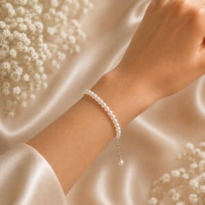 Puede incluir: Una delicada pulsera de perlas con un peque&ntilde;o dije de perla colgante. La pulsera est&aacute; compuesta por peque&ntilde;as perlas blancas redondas ensartadas con una cadena de plata. La pulsera se exhibe sobre una superficie de sat&eacute;n color crema.