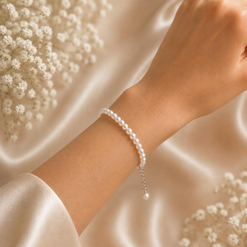 Puede incluir: Una delicada pulsera de perlas con un peque&ntilde;o dije de perla colgante. La pulsera est&aacute; compuesta por peque&ntilde;as perlas blancas redondas ensartadas con una cadena de plata. La pulsera se exhibe sobre una superficie de sat&eacute;n color crema.