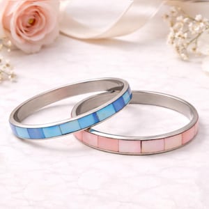 May include: Two silver bangles. One bangle features a mosaic of light and dark blue squares. The other bangle has a mosaic of pink squares. The background includes a pink rose and white ribbon.