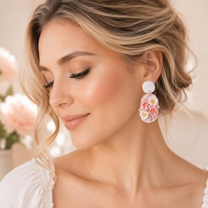 Puede incluir: Un par de pendientes colgantes con un poste circular blanco conectado a un colgante ovalado. El colgante es de color rosa claro y está decorado con pequeñas flores esculpidas en tres dimensiones en tonos blanco, rosa y amarillo.