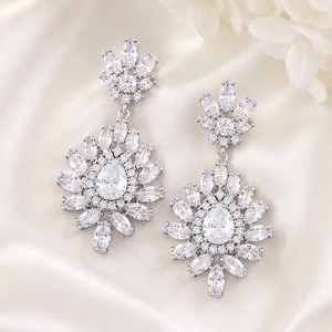 May include: A pair of silver-toned drop earrings featuring a cluster of clear, faceted marquise and round-cut stones. Each earring has a top floral-shaped component connected to a larger, circular pendant with a central pear-shaped stone.