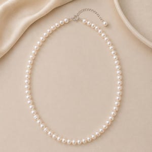 Puede incluir: Un delicado collar de perlas con un cierre plateado. El collar presenta una hebra de perlas redondas de color blanco cremoso. El cierre es de dise&ntilde;o sencillo y elegante. El collar se exhibe sobre un fondo neutro.