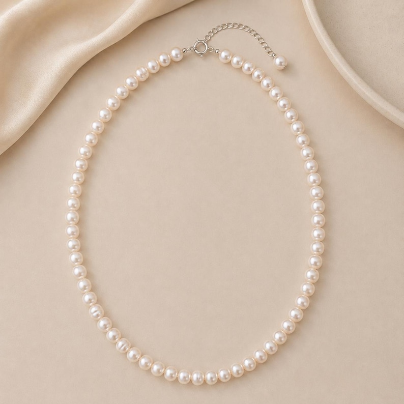 Puede incluir: Un delicado collar de perlas con un cierre plateado. El collar presenta una hebra de perlas redondas de color blanco cremoso. El cierre es de dise&ntilde;o sencillo y elegante. El collar se exhibe sobre un fondo neutro.