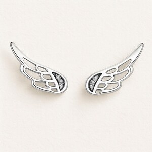 May include: A pair of silver-toned wing-shaped earrings. Each earring features an openwork design with a curved shape and small, sparkling stones set near the base. The earrings are displayed against a soft, off-white background.