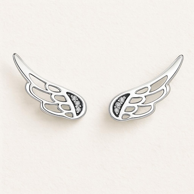 May include: A pair of silver-toned wing-shaped earrings. Each earring features an openwork design with a curved shape and small, sparkling stones set near the base. The earrings are displayed against a soft, off-white background.