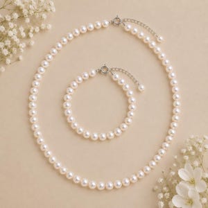 Puede incluir: Un conjunto de collar y pulsera de perlas. La joyer&iacute;a presenta perlas redondas y blancas ensartadas con un cierre plateado. El collar es m&aacute;s largo, mientras que la pulsera es m&aacute;s corta. El conjunto se exhibe sobre una superficie beige con flores blancas.