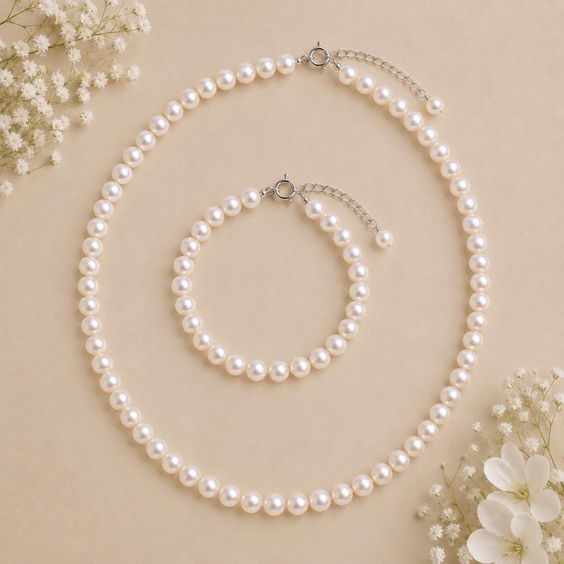 Puede incluir: Un conjunto de collar y pulsera de perlas. La joyer&iacute;a presenta perlas redondas y blancas ensartadas con un cierre plateado. El collar es m&aacute;s largo, mientras que la pulsera es m&aacute;s corta. El conjunto se exhibe sobre una superficie beige con flores blancas.