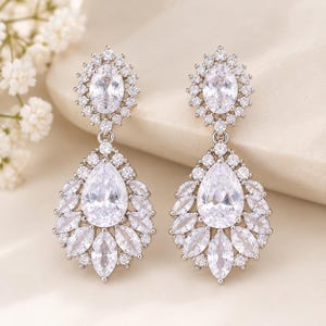 May include: A pair of silver-toned drop earrings featuring a central oval-cut clear stone surrounded by a halo of small round stones, connected to a larger pear-shaped drop base adorned with marquise and round-cut clear stones in a cluster.