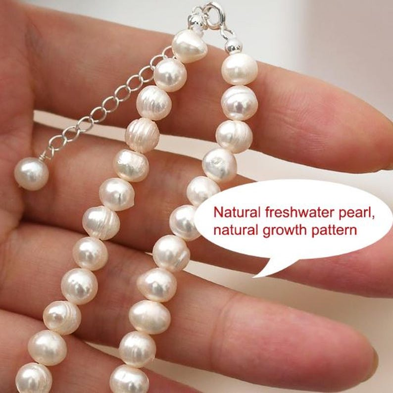 Puede incluir: Primer plano de un collar de perlas con cadena y cierre plateados. El collar est&aacute; hecho de perlas blancas y redondas. El texto de la imagen dice: "Perla natural de agua dulce, patr&oacute;n de crecimiento natural."