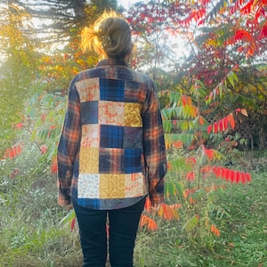 Può includere: Una camicia a maniche lunghe con patchwork con un motivo a quadri arancione, blu e marrone. La parte posteriore della camicia presenta una varietà di quadrati di tessuto con diversi motivi e colori. La persona indossa pantaloni neri ed è in un ambiente naturale.