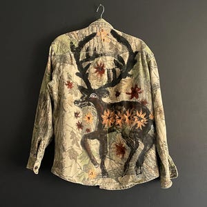 以下が含まれることがあります： 背中に大きな鹿のイラストが描かれた迷彩柄のボタンダウンシャツ。鹿は茶色で、黒い角とオレンジと赤の花柄のアクセントが付いています。シャツは長袖で、ボタンダウンフロントです。