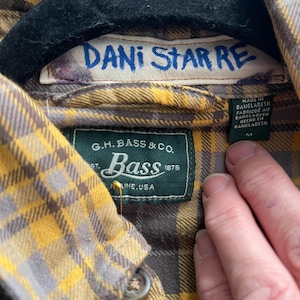 Op de afbeelding: Close-up van een geel en grijs geruit overhemd. Het label leest "DANI STAR RE" en "G.H. Bass & Co.". Het overhemd is gemaakt in Bangladesh. De kraag is geknoopt.