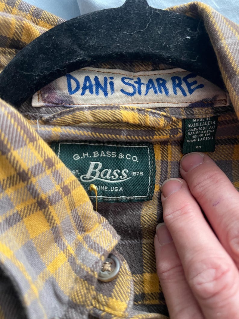 Op de afbeelding: Close-up van een geel en grijs geruit overhemd. Het label leest "DANI STAR RE" en "G.H. Bass & Co.". Het overhemd is gemaakt in Bangladesh. De kraag is geknoopt.