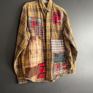 Op de afbeelding: Een overhemd met knoopsluiting en een patchwork-ontwerp. Het overhemd heeft een ruitpatroon in bruin-, geel- en roodtinten. Verschillende stoffen patches in verschillende patronen en kleuren zijn op het overhemd genaaid, wat een unieke uitstraling cre&euml;ert.