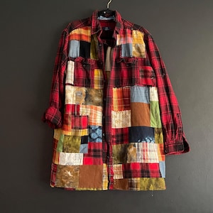 May include: A long-sleeved, patchwork shirt in a red plaid pattern with various fabric squares in shades of red, yellow, blue, and brown. The shirt is buttoned and hangs on a black hanger against a dark wall.