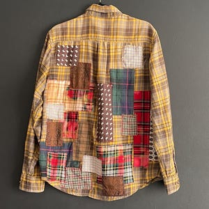 May include: A button-down shirt with a yellow and brown plaid pattern. The back of the shirt is adorned with various patches of different fabrics and patterns, including plaid, floral, and solid colors. The shirt is hanging on a black hanger.