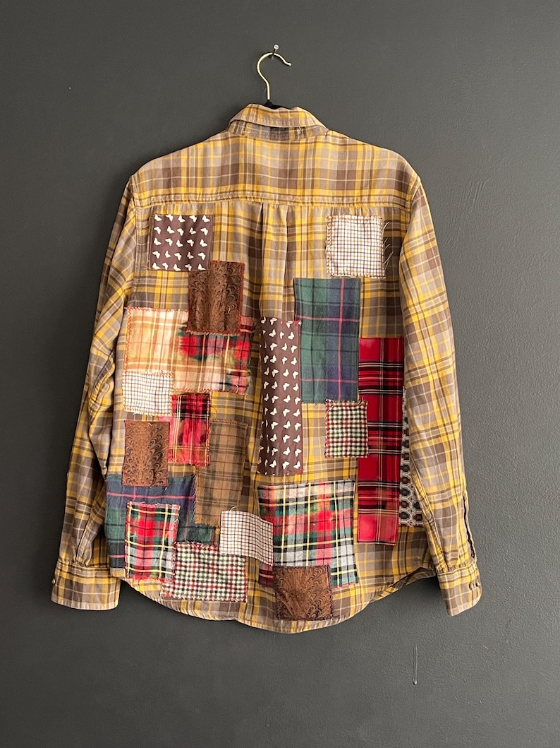 Op de afbeelding: Een button-down overhemd met een geel en bruin geruit patroon. De achterkant van het overhemd is versierd met verschillende patches van verschillende stoffen en patronen, waaronder ruitjes, bloemen en effen kleuren. Het overhemd hangt aan een zwarte hanger.