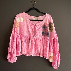 Op de afbeelding: Een roze top met lange mouwen en een gerimpelde taille en mouwen. De top heeft verschillende patches van verschillende stoffen, waaronder ruit- en bloemmotieven. Het hangt aan een zwarte hanger tegen een donkergrijze achtergrond.