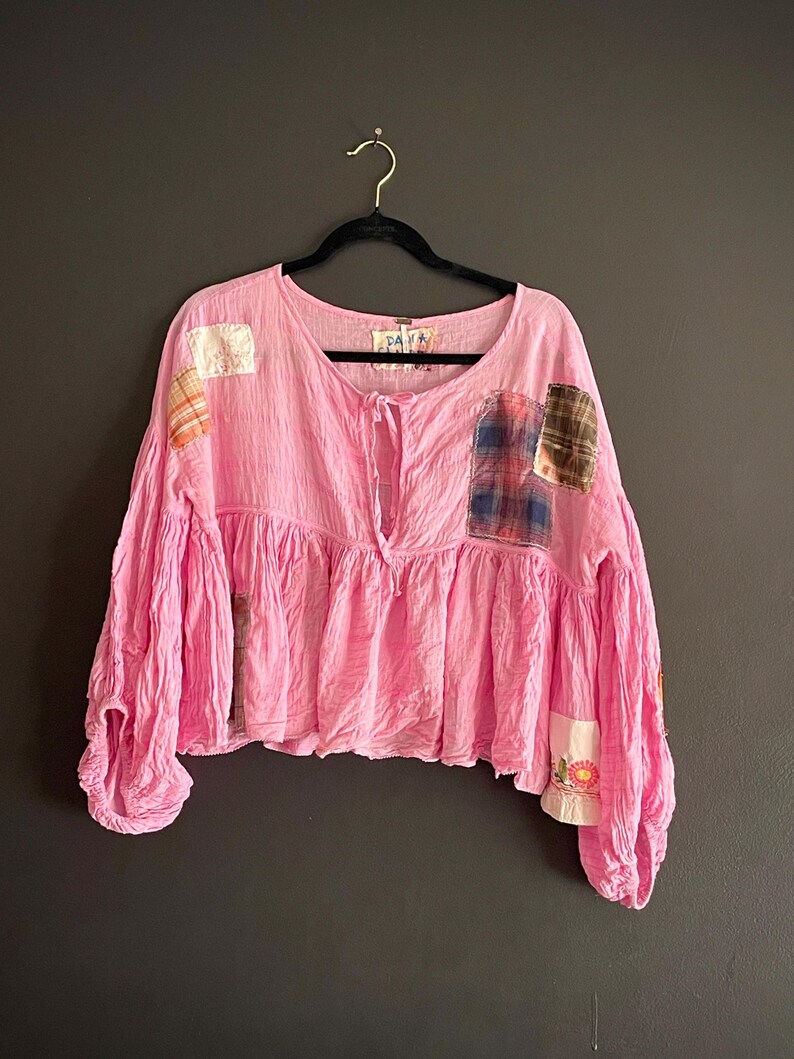 Op de afbeelding: Een roze top met lange mouwen en een gerimpelde taille en mouwen. De top heeft verschillende patches van verschillende stoffen, waaronder ruit- en bloemmotieven. Het hangt aan een zwarte hanger tegen een donkergrijze achtergrond.