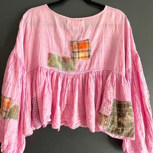 Op de afbeelding: Een roze top met ronde hals en patchwork details. De top heeft een geplooid, gerimpeld tailleontwerp en verschillende geruite patches in oranje, groen en bruin. De mouwen zijn volumineus.