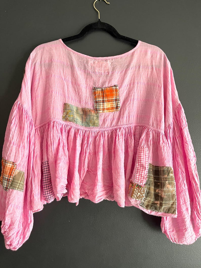 Op de afbeelding: Een roze top met ronde hals en patchwork details. De top heeft een geplooid, gerimpeld tailleontwerp en verschillende geruite patches in oranje, groen en bruin. De mouwen zijn volumineus.