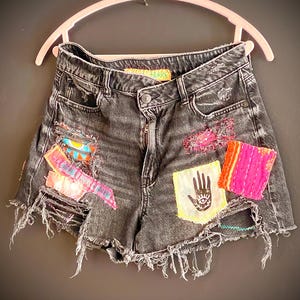 May include: Distressed black denim shorts with colorful patches and frayed edges. Patches include a hand with an eye design, and other colorful fabric pieces. The shorts are hanging on a pink hanger.