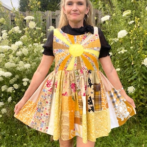 May include: A sleeveless patchwork dress featuring a yellow sun applique on the bodice with radiating rays. The skirt is constructed from various yellow, gold, and white fabric squares with floral, plaid, and animal patterns in a flared silhouette.