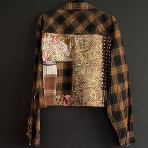 May include: A long-sleeve button-up shirt featuring a brown and black plaid pattern on the collar, sleeves, and upper back. The back panel is a patchwork design with floral, toile, and geometric fabric squares in brown, tan, and white tones.