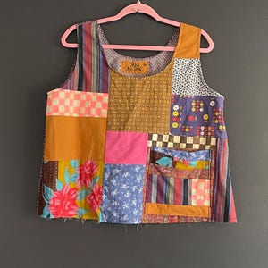 May include: A sleeveless patchwork top featuring a variety of fabric patterns including floral, checkered, striped, and solid mustard yellow panels. The garment has a square neckline, a front pocket, and a label reading DANI STARRE at the collar.