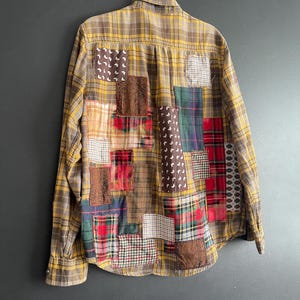 Op de afbeelding: Een button-down shirt met lange mouwen en een geel-bruin geruit patroon. De achterkant van het shirt is versierd met verschillende patches van verschillende stoffen, waaronder ruitjes, bloemen en textuurmaterialen. Het shirt hangt aan een metalen hanger.